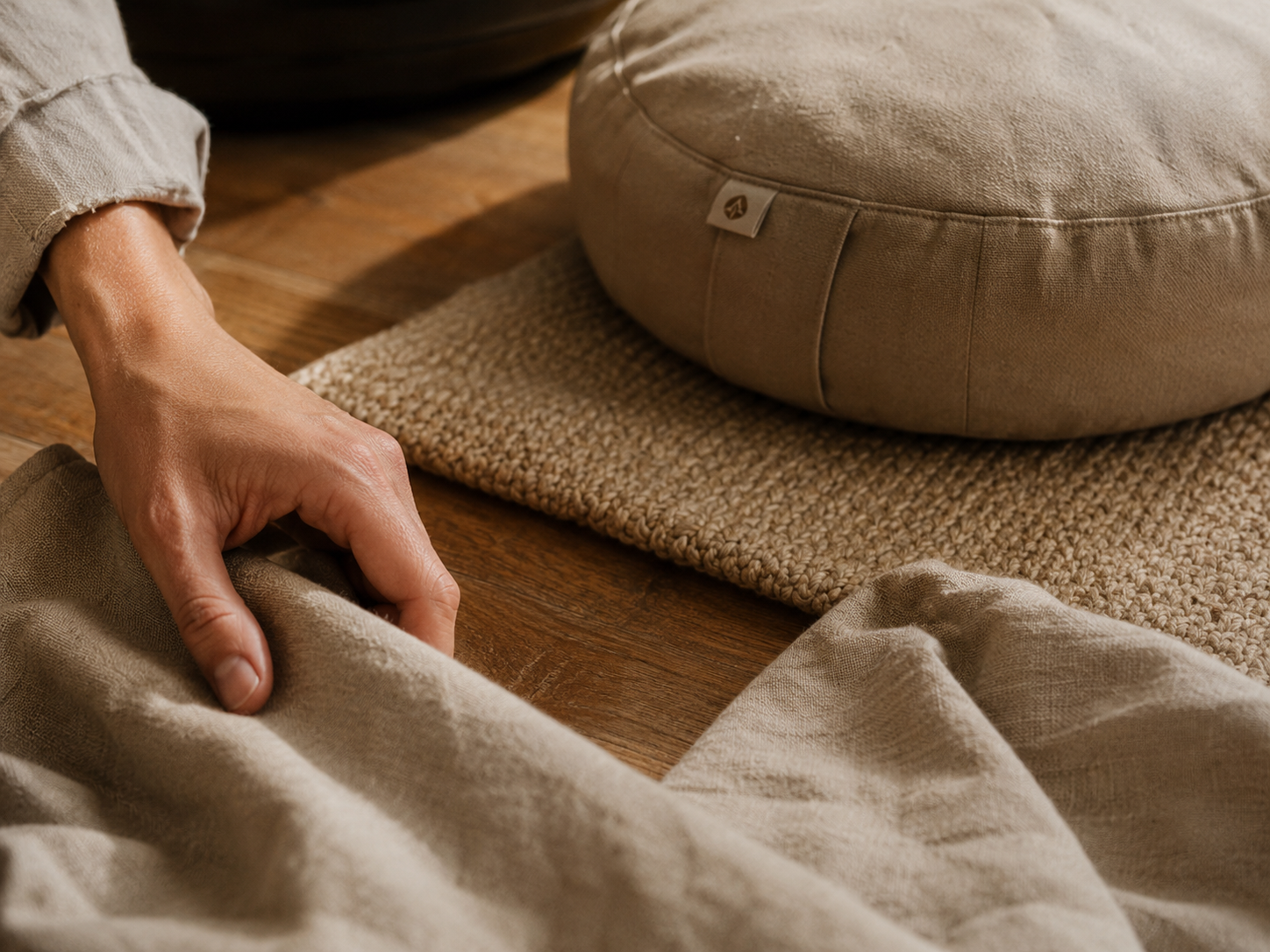 Hand touching natural linen beside a meditation cushion on a woven mat in warm natural light.
